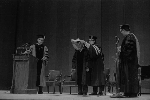 Conferral of an honorary doctoral degree on Shinichi Suzuki. Professor Donald Shetler hoods the newly named Dr. Suzuki; University President Robert Sproull stands at far left, and ESM Director Walter Hendl stands at far right.